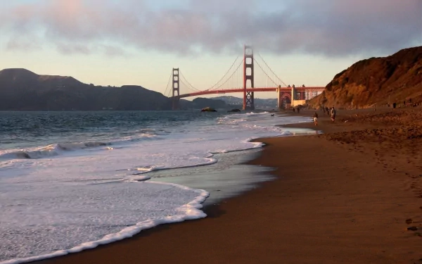 HD desktop wallpaper featuring the man-made Golden Gate Bridge viewed from a sandy beach at sunset with waves gently washing ashore.