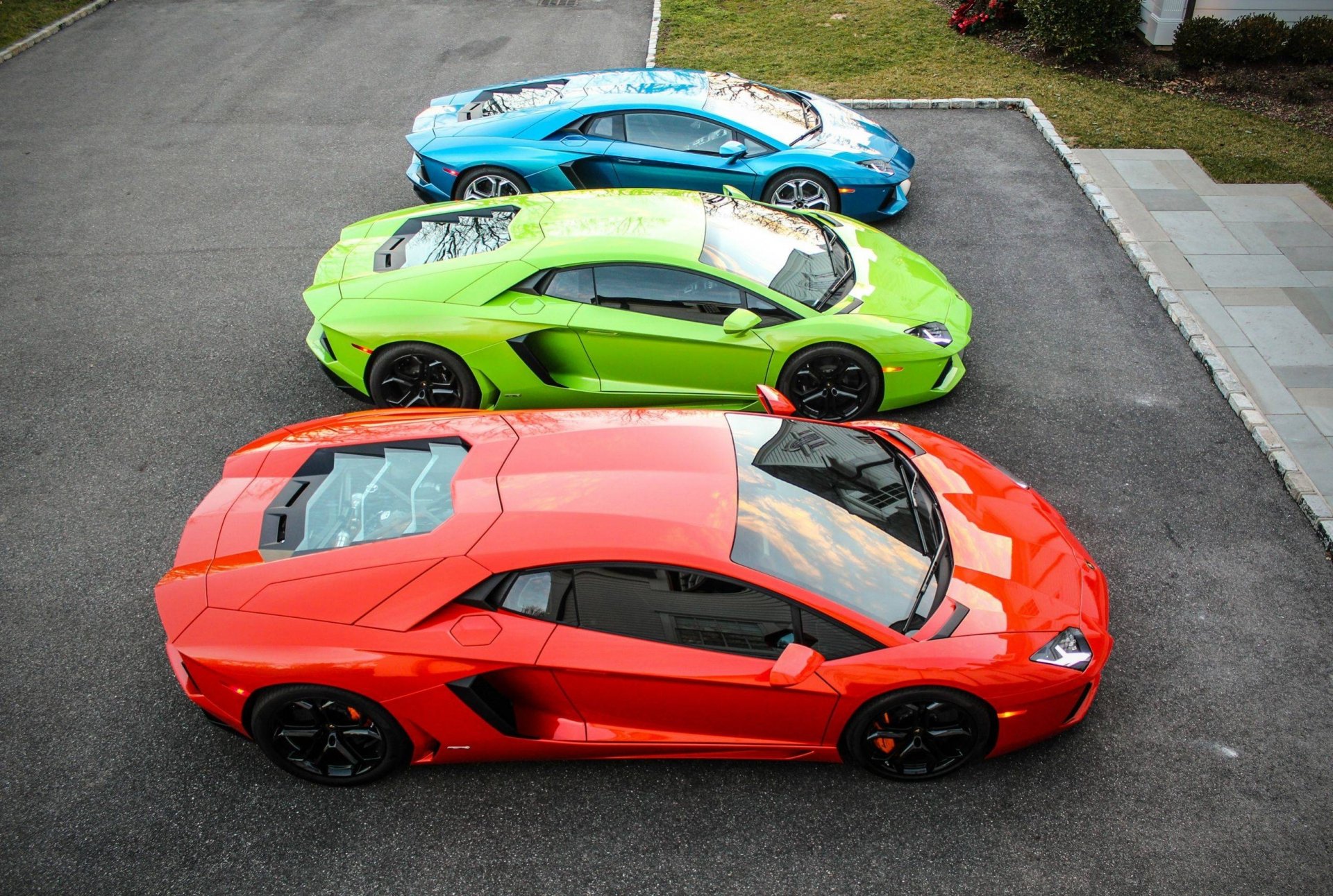 A stunning HD wallpaper featuring a trio of Lamborghini Aventadors in vibrant colors: orange, green, and blue, showcased from an aerial perspective on a sleek driveway.