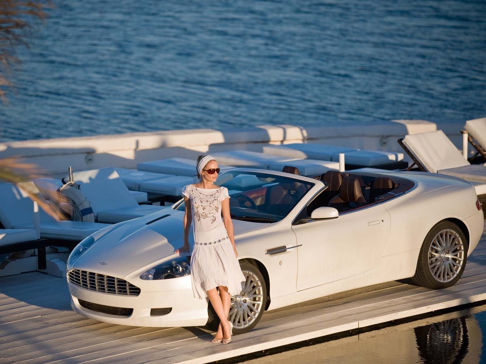 HD PC desktop wallpaper: white Aston Martin DB9 convertible on a marina dock, a woman in a white dress leaning against the car with blue ocean in the background.