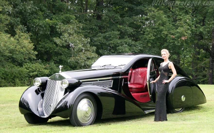 A sleek black 1925 Rolls-Royce Phantom with a red interior showcased on a grassy lawn, accompanied by a woman in an elegant black dress.