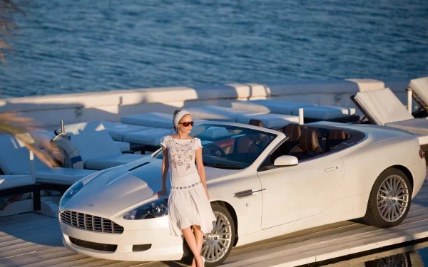HD PC desktop wallpaper: white Aston Martin DB9 convertible on a marina dock, a woman in a white dress leaning against the car with blue ocean in the background.