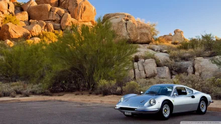 A silver Ferrari Dino parked on a desert road with rocky hills and green shrubs under a clear blue sky, captured in a high-definition PC desktop wallpaper.