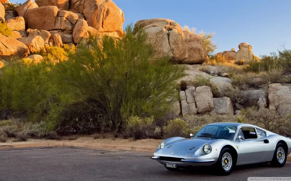 A silver Ferrari Dino parked on a desert road with rocky hills and green shrubs under a clear blue sky, captured in a high-definition PC desktop wallpaper.