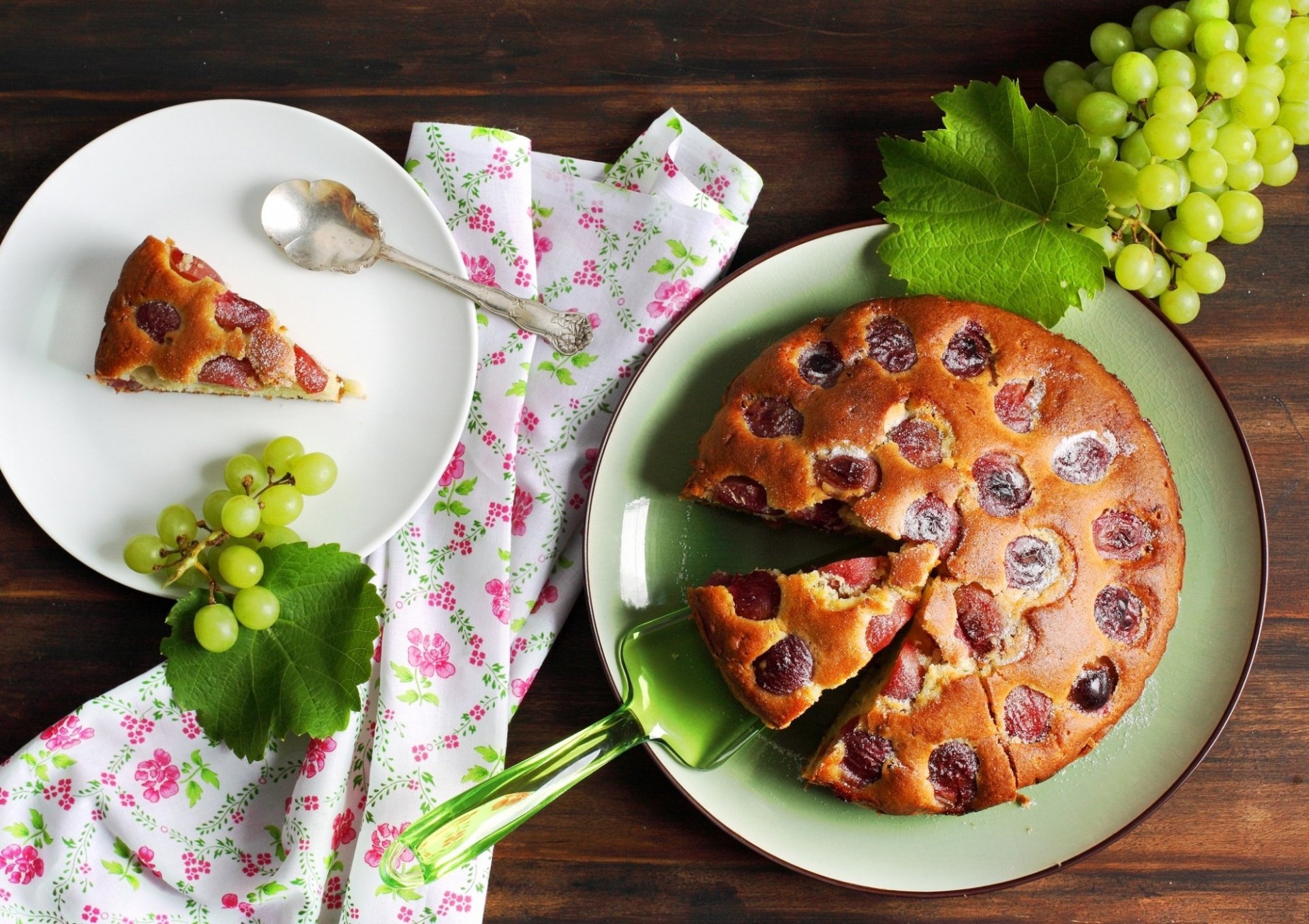 HD desktop wallpaper featuring a freshly baked cherry cake with slices served on plates, accompanied by green grapes and decorative leaves on a wooden table.
