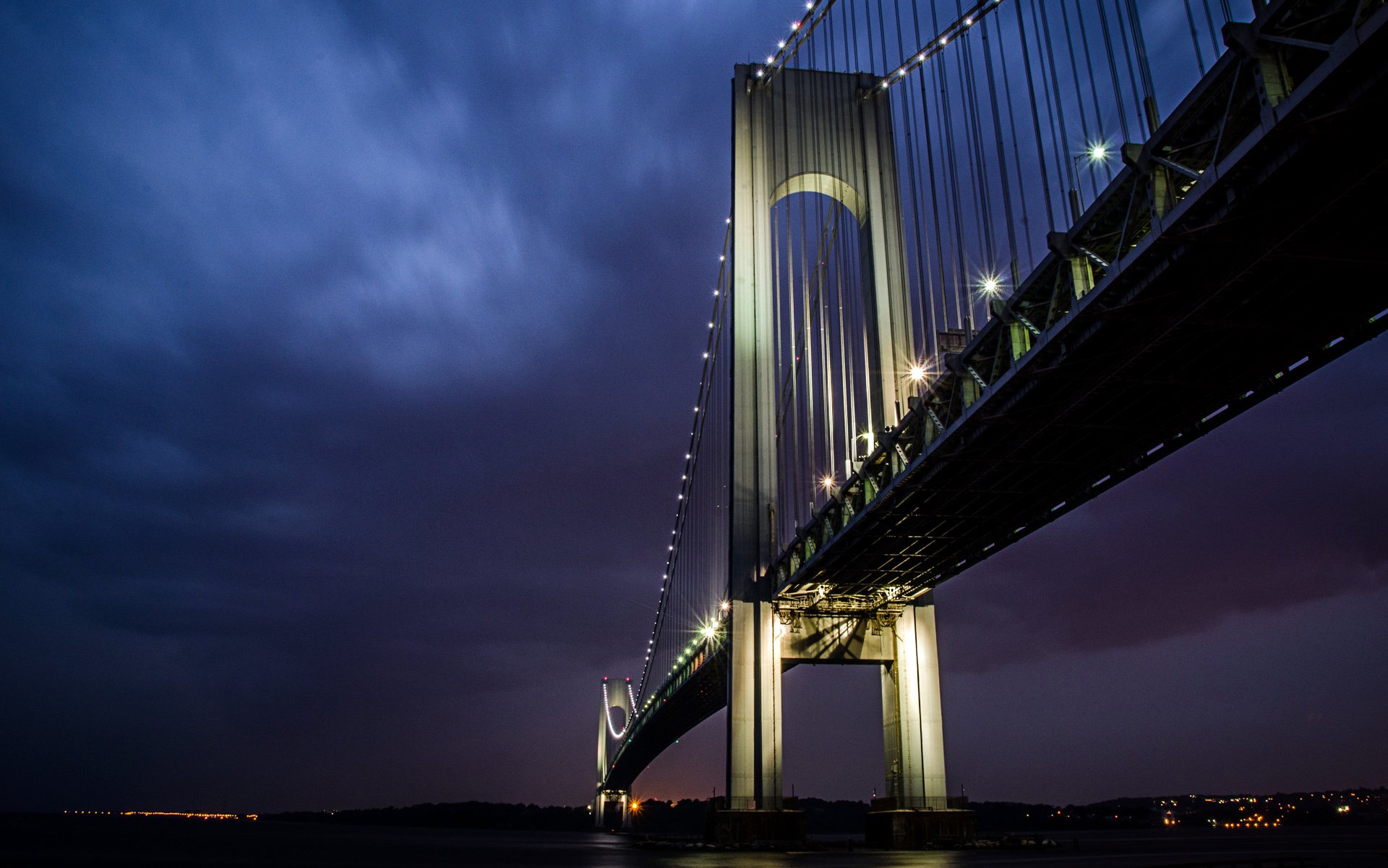 Night view of the illuminated Verrazzano-Narrows Bridge in New York, showcasing its man-made structure against a dark, cloudy sky, captured in HD.