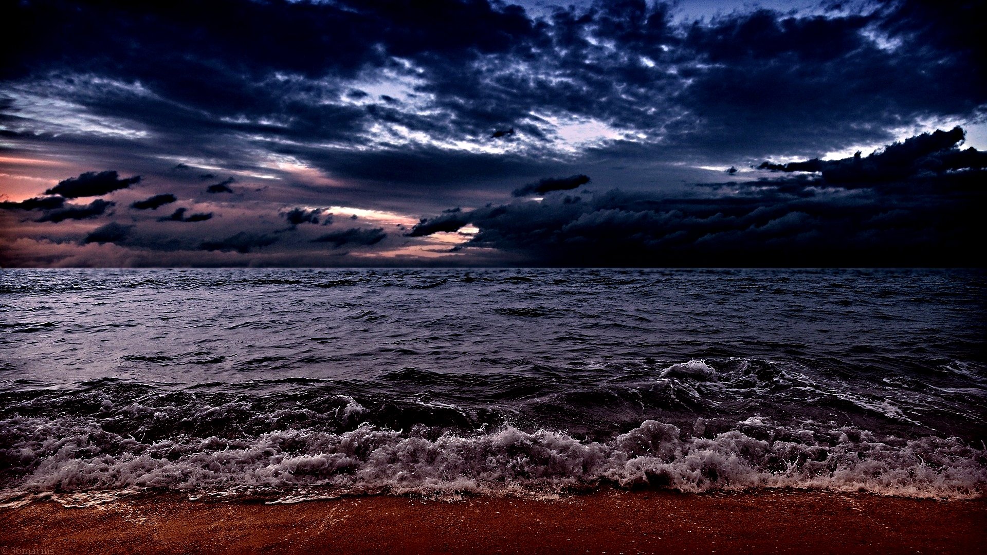 HD desktop wallpaper showing a dramatic ocean scene at dusk with waves crashing onto the shore under a cloudy, darkening sky.
