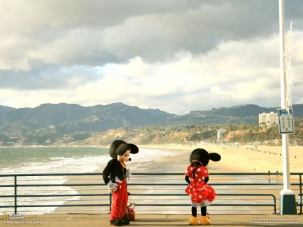 Funny cartoon HD PC desktop wallpaper and background: two cartoon mice—one in a red polka-dot dress—chat on a pier overlooking a sunlit beach, ocean and distant mountains.