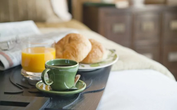 A still life HD desktop wallpaper featuring a breakfast scene with a cup of coffee, glass of juice, and croissants on a tray by the bedside.