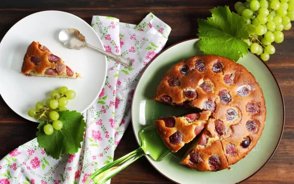 HD desktop wallpaper featuring a freshly baked cherry cake with slices served on plates, accompanied by green grapes and decorative leaves on a wooden table.