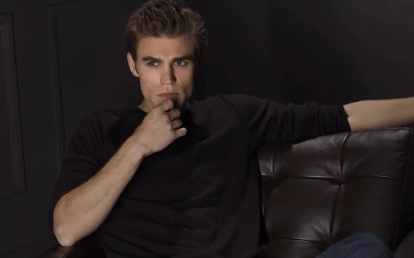 HD desktop wallpaper featuring celebrity Paul Wesley seated on a dark leather couch against a black backdrop, dressed in a black long-sleeve shirt.