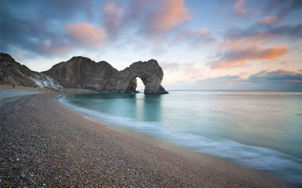 HD PC desktop wallpaper of Durdle Door: a natural limestone cliff arch on a pebbled beach with calm turquoise sea and a pastel sunset sky.