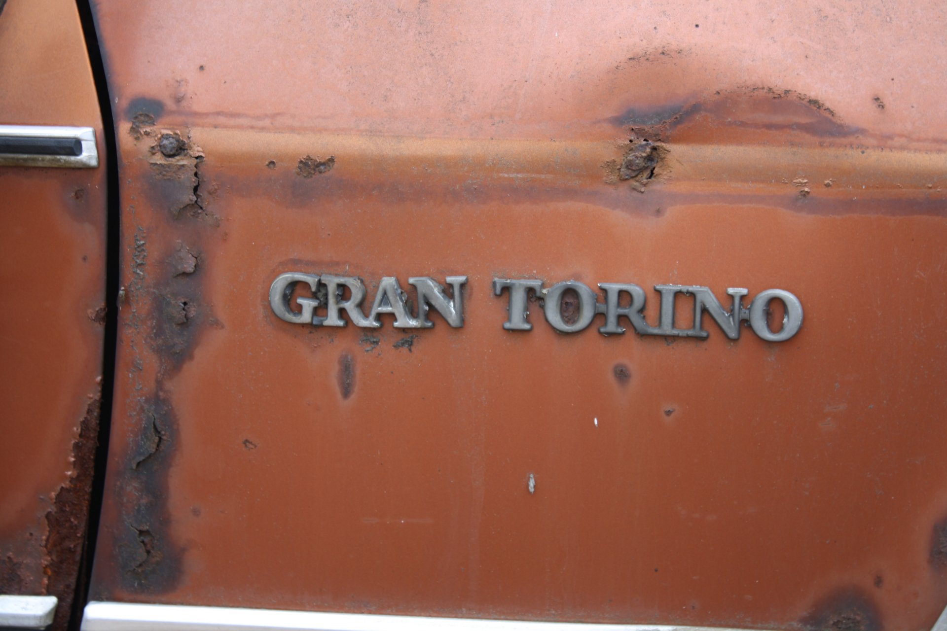 Close-up of a rusty, weathered Ford Gran Torino emblem on the orange door panel, captured in 4K Ultra HD for a PC desktop wallpaper.