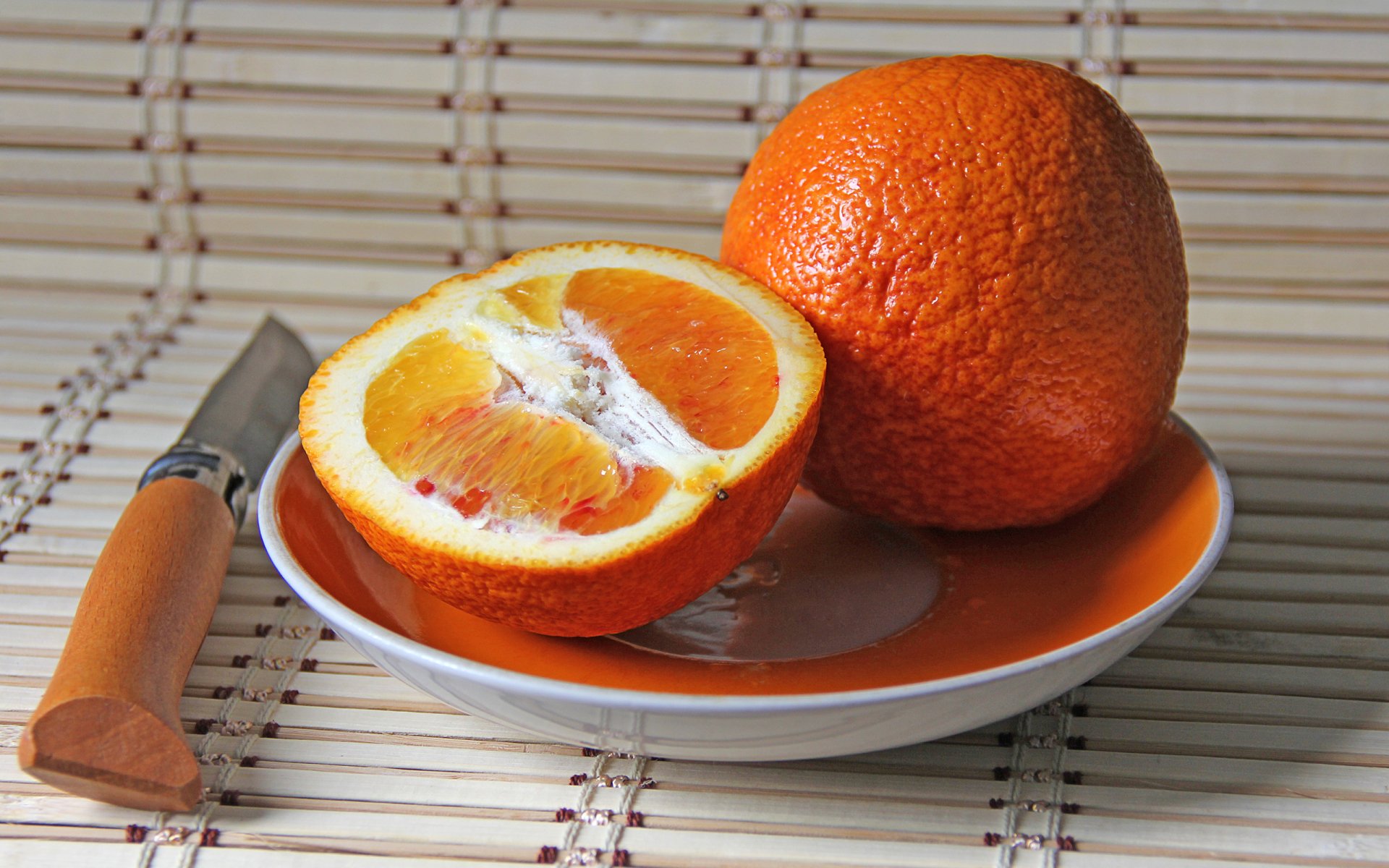 HD desktop wallpaper featuring a vibrant orange fruit, whole and halved, resting on an orange and white plate with a knife beside it on a bamboo mat.