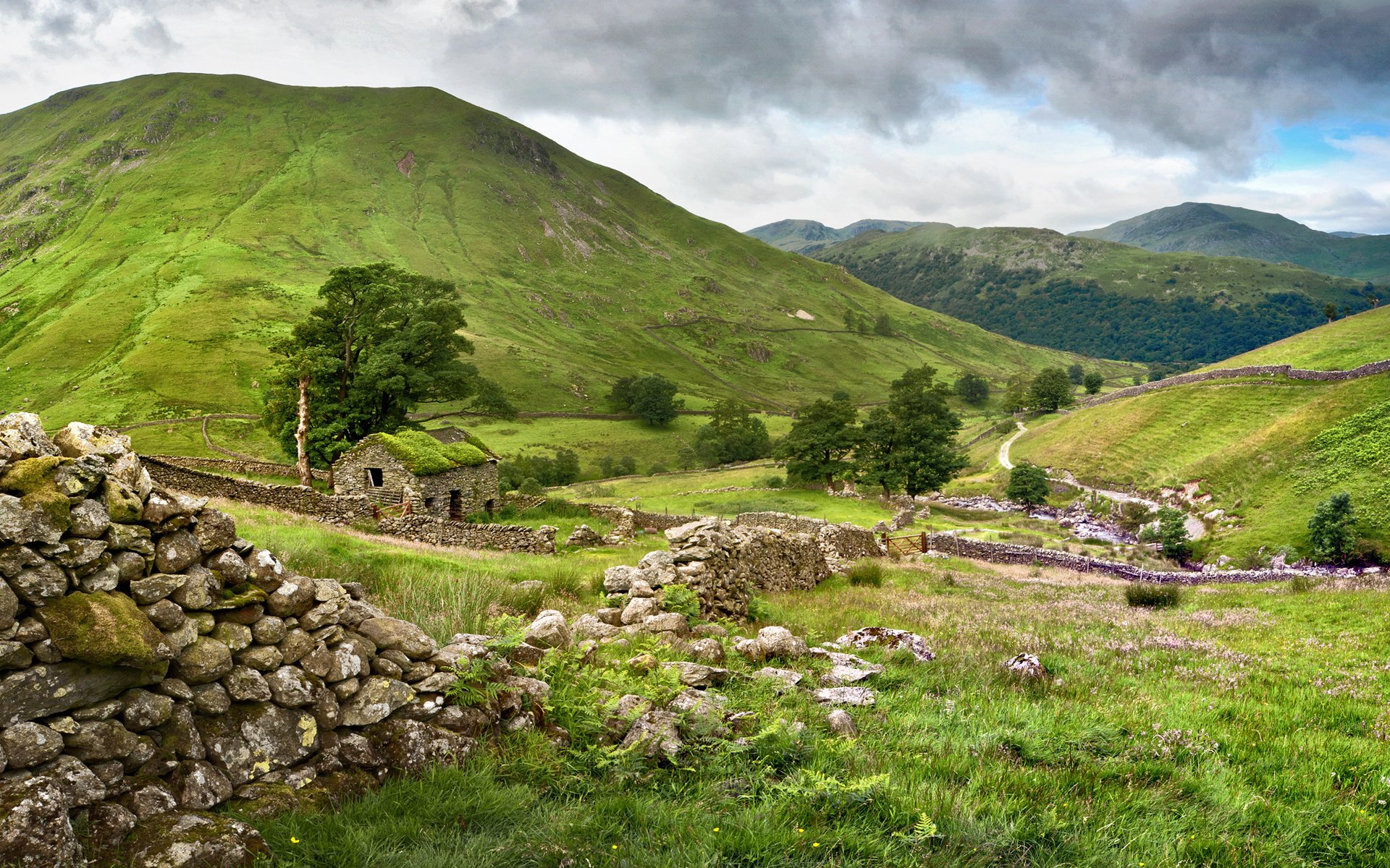 HD desktop wallpaper of a man-made stone cabin nestled in lush green hills under a cloudy sky, surrounded by stone walls and vibrant natural scenery.