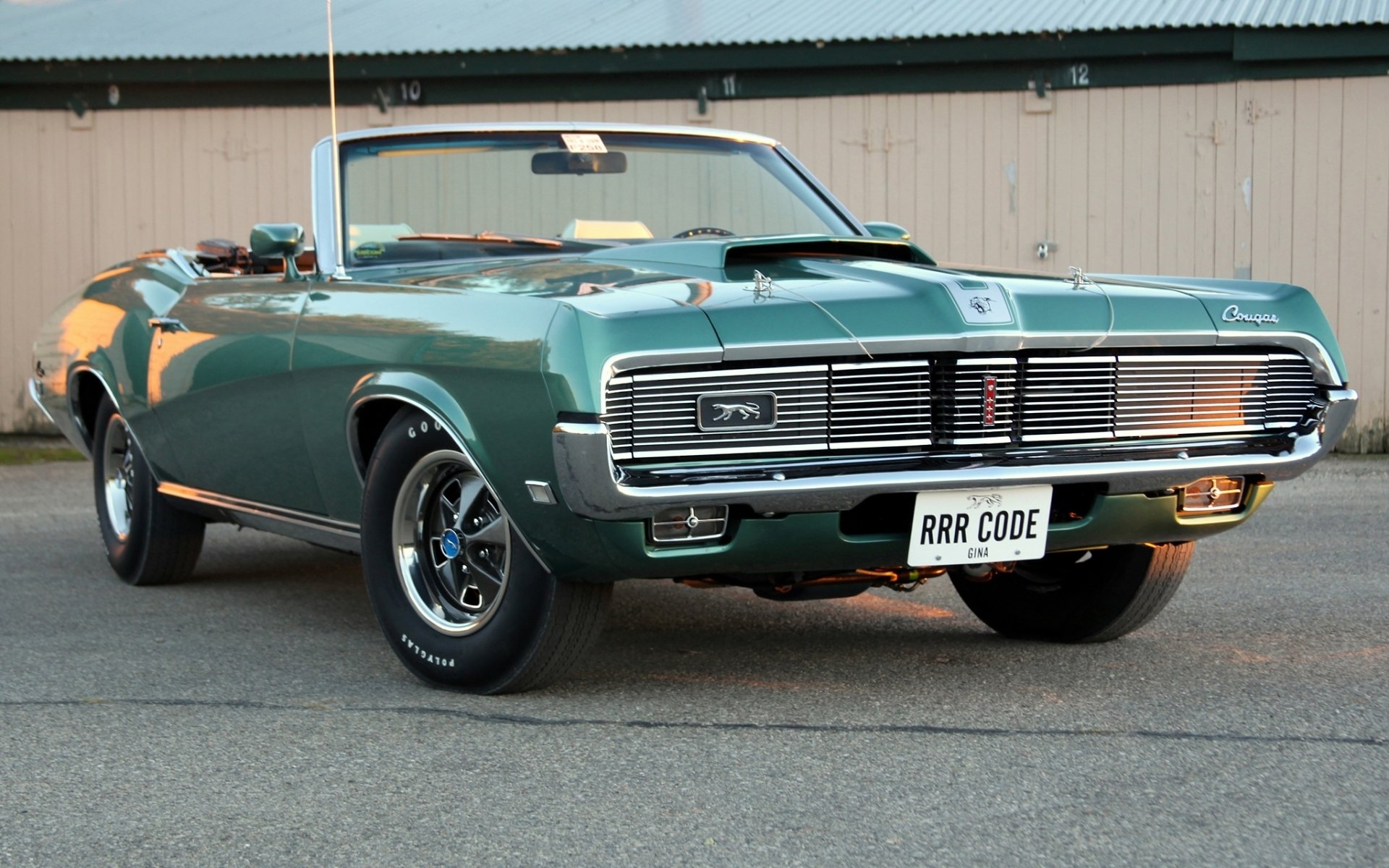 HD desktop wallpaper featuring a green 1969 Mercury Cougar convertible parked on pavement against a beige industrial backdrop.