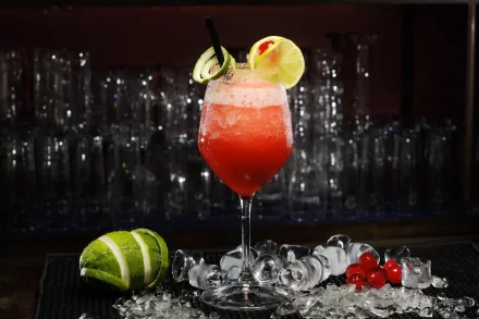 A vibrant cocktail garnished with lime and cherry, served over ice, stands against a blurred background of glassware, capturing the essence of refreshing indulgence. 4K Ultra HD desktop wallpaper.