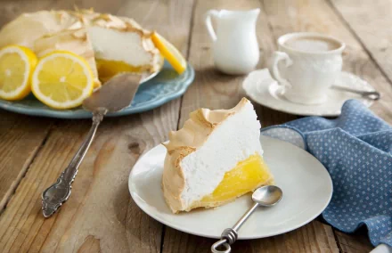 A slice of lemon meringue pie on a delicate plate, accompanied by fresh lemons, a cup of tea, and a blue patterned napkin, set on a rustic wooden table. An inviting food-themed wallpaper.