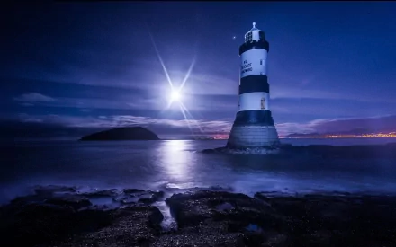 A striking HD wallpaper featuring a man-made lighthouse standing tall against a twilight sky, with the moon reflecting on the ocean waves and distant land visible.