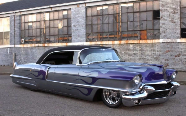 HD PC desktop wallpaper of a classic Cadillac with a custom blue flame paint job, parked in front of an industrial brick building with large windows.