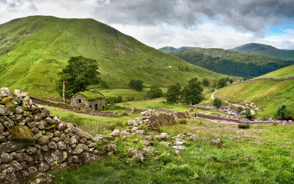 HD desktop wallpaper of a man-made stone cabin nestled in lush green hills under a cloudy sky, surrounded by stone walls and vibrant natural scenery.