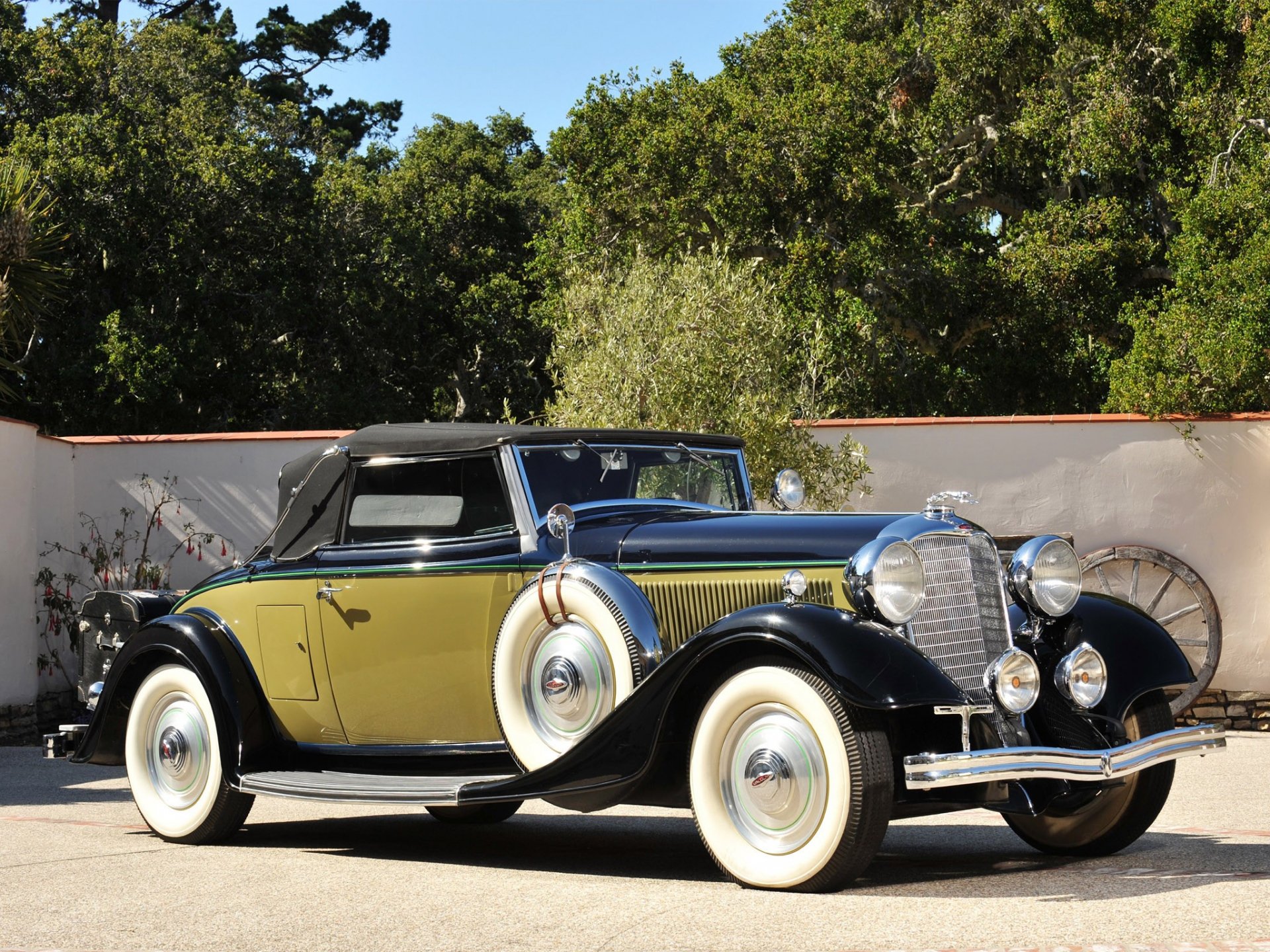 HD desktop wallpaper featuring a classic 1933 Lincoln Model KA vehicle in black and yellow, parked outdoors with trees and a wall in the background.