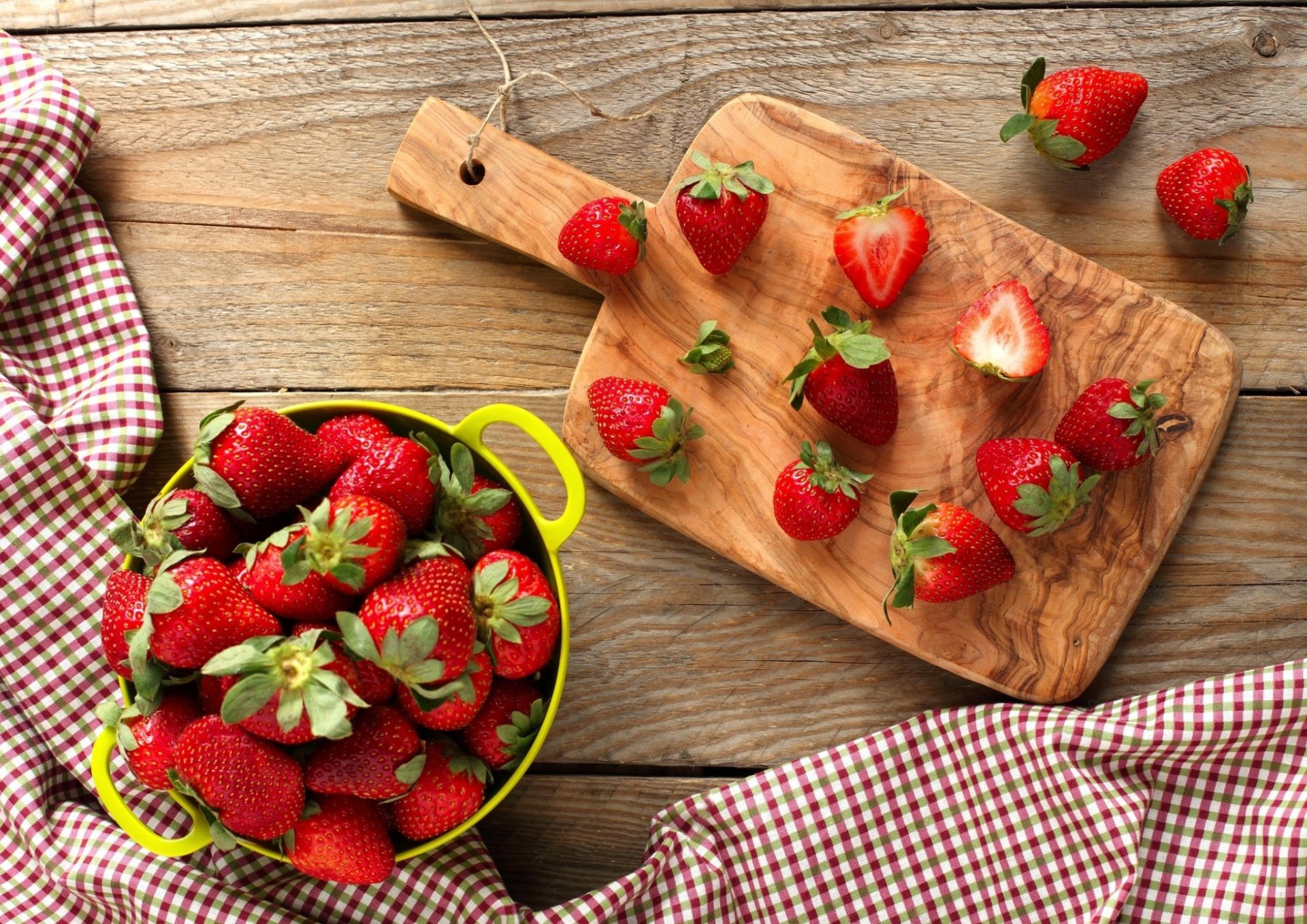 HD PC desktop wallpaper featuring fresh strawberries on a wooden cutting board and in a green colander, set on a rustic wooden table with a checkered cloth.