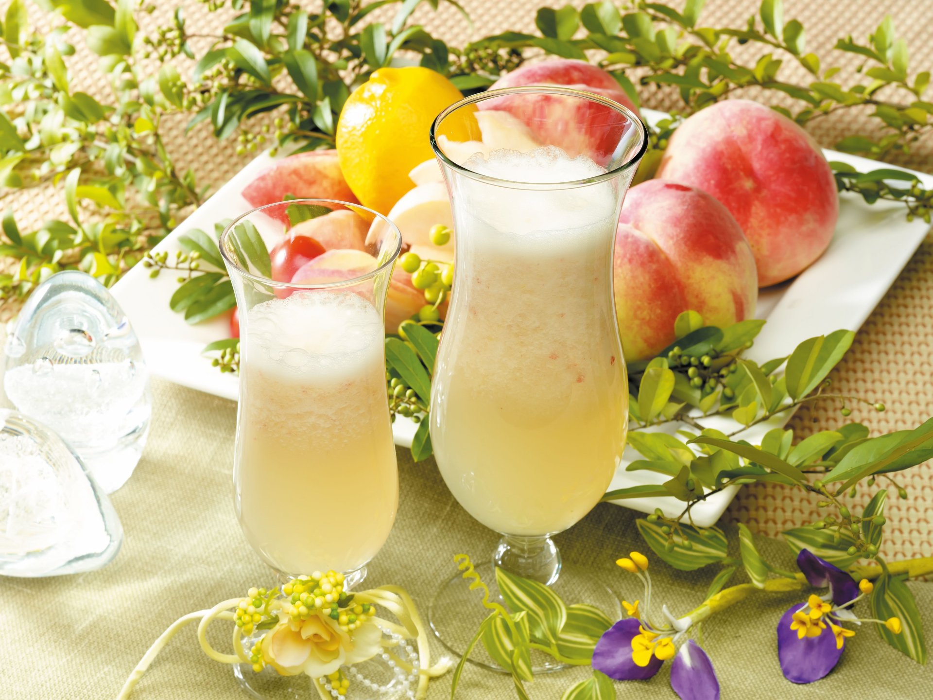 4K Ultra HD desktop wallpaper featuring two creamy peach cocktails surrounded by fresh peaches, flowers, and ice on a natural woven surface.