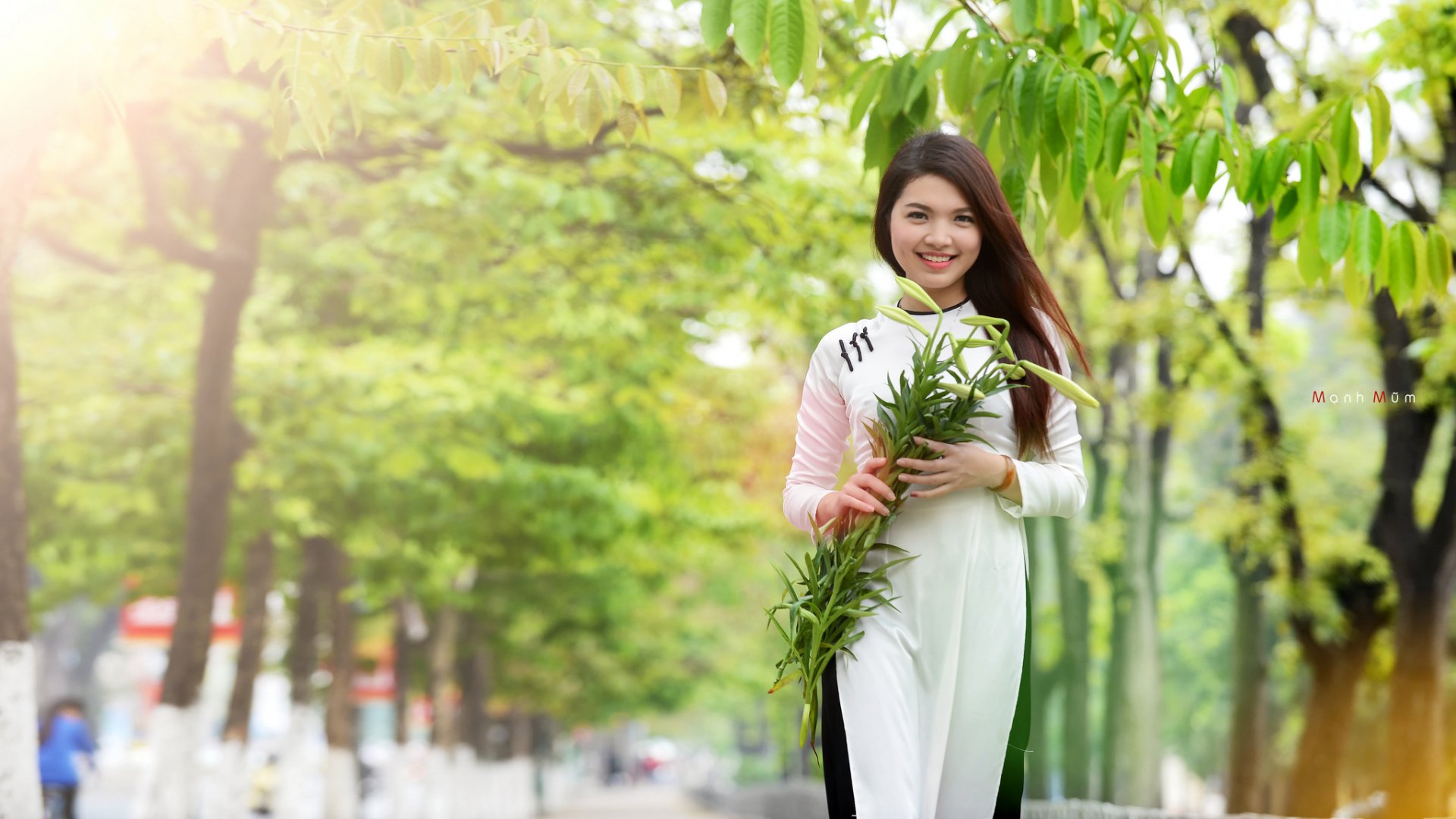 A smiling Asian woman in a traditional outfit holds greenery, walking through a lush, tree-lined path. This vibrant HD image serves as an engaging desktop wallpaper.