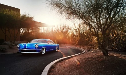 A vibrant blue 1951 Kaiser Dragon Coupe lowrider classic car parked on a winding road at sunset, showcasing custom Chevrolet styling in a scenic desert setting.