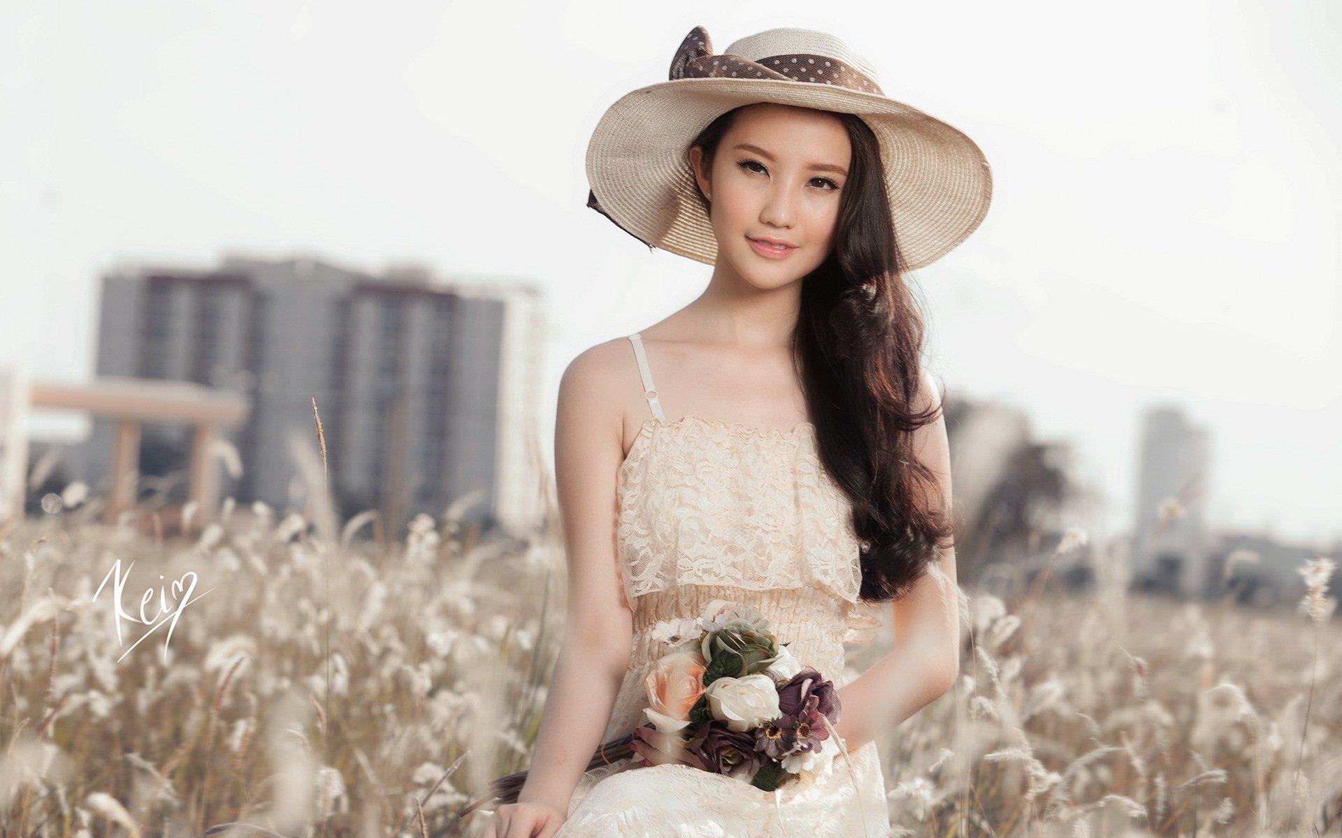 An Asian woman in a white dress and hat stands in a field of tall grasses, holding flowers, with buildings blurred in the background. HD desktop wallpaper and background.