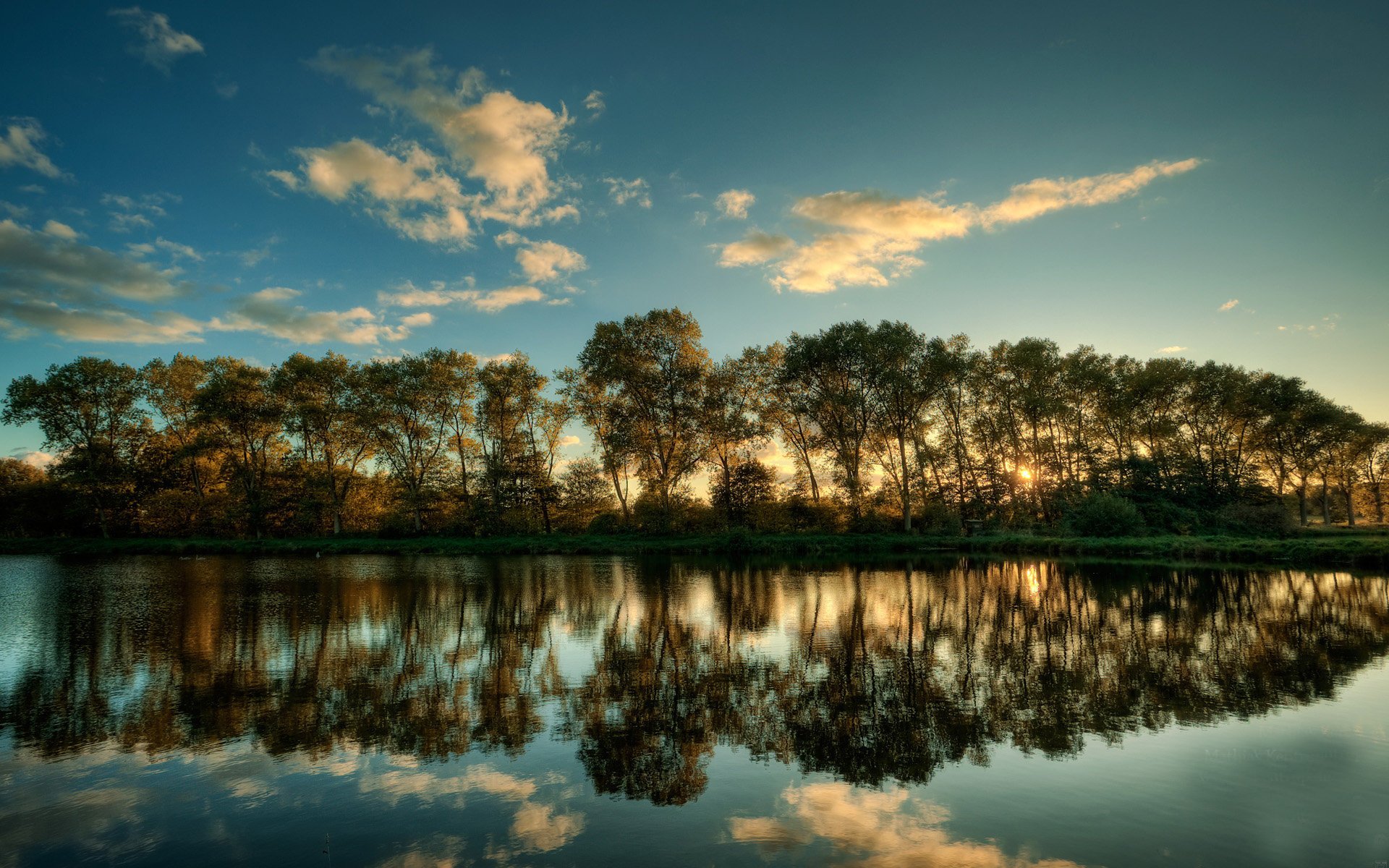 HD PC desktop wallpaper background: nature scene of a tree-lined shoreline at sunset, reflected in a calm lake beneath a streaked blue sky.