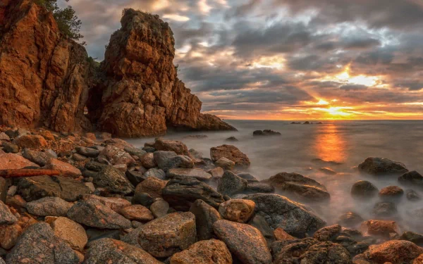HD PC desktop wallpaper featuring a rocky coastline at sunset with dramatic clouds and soft waves, showcasing the beauty of nature.