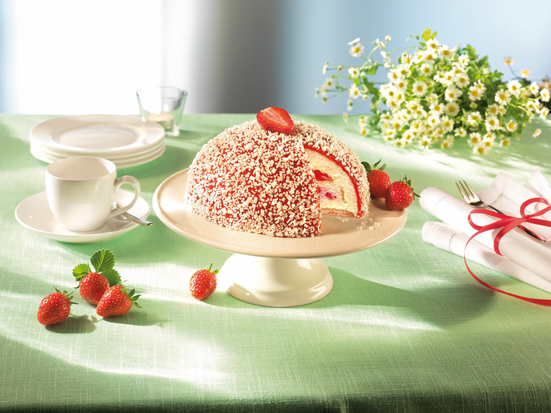 A delectable cake adorned with strawberries and chocolate sprinkles, surrounded by a chamomile bouquet, dessert plates, and a cup, set against a soft green background.