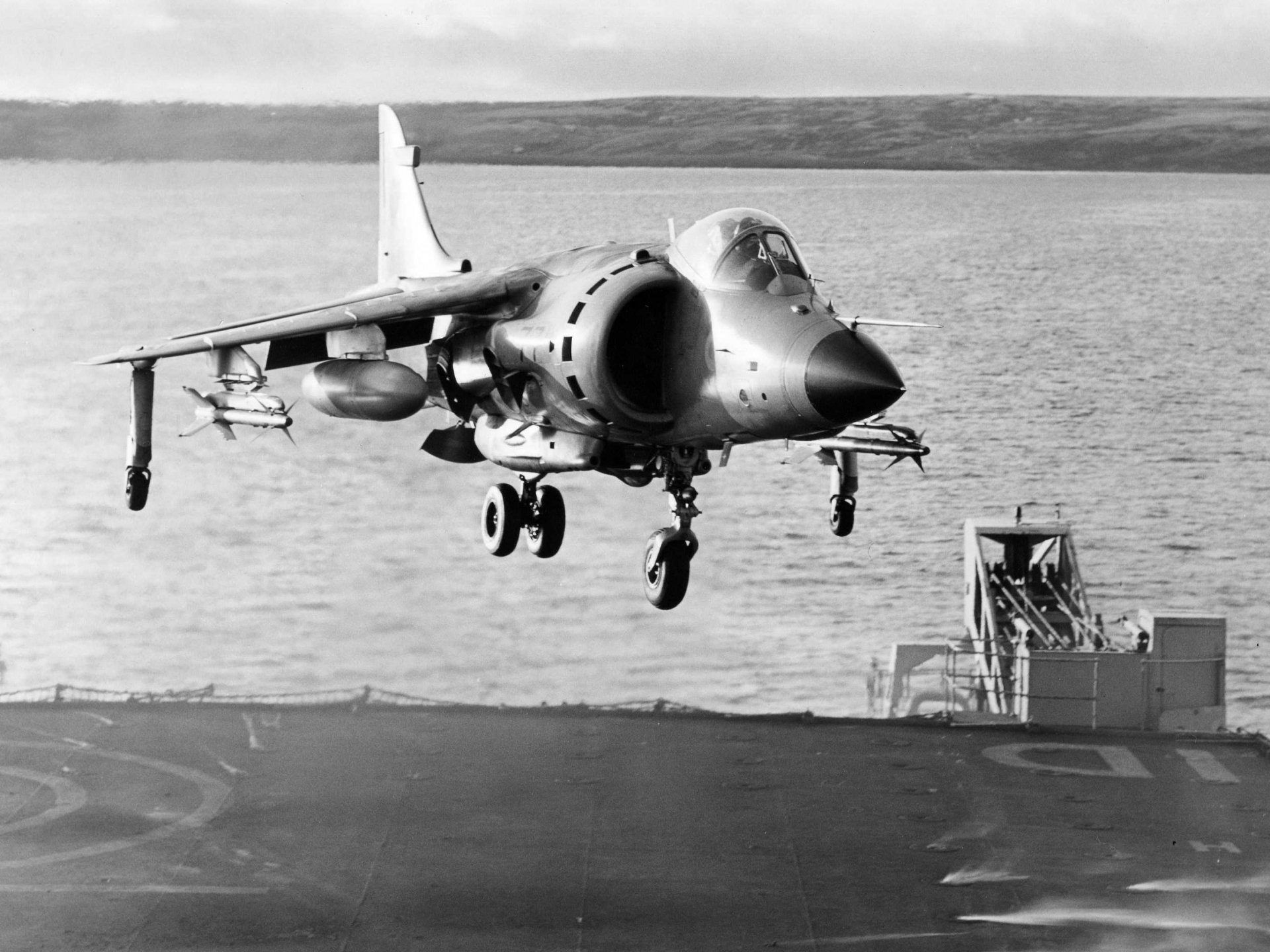 Black and white image of a McDonnell Douglas AV-8B Harrier II military jet hovering over a naval ship, captured as an HD desktop wallpaper and background.