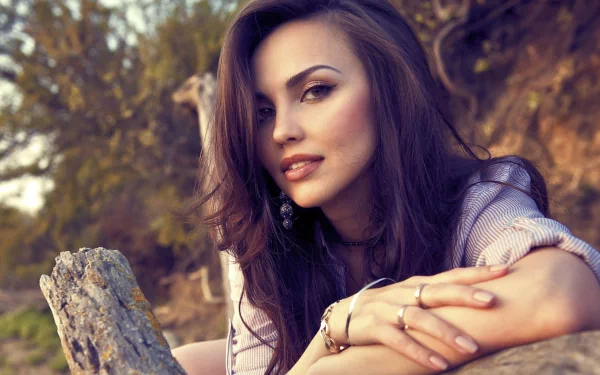 HD wallpaper featuring a beautiful woman with long dark hair, lounging on a wooden surface with a natural background of trees and shrubs. She gazes confidently at the camera.