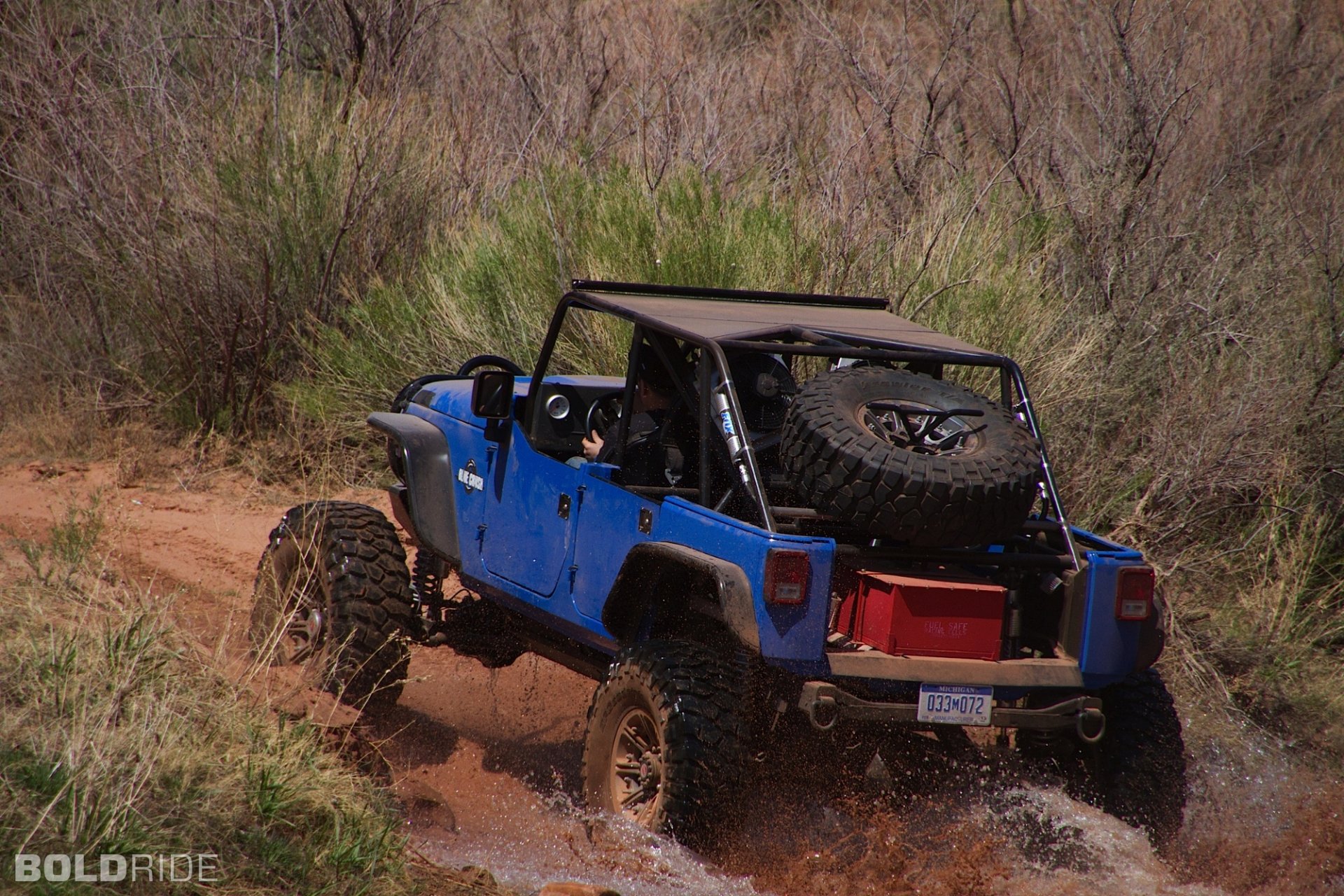 HD PC desktop wallpaper featuring a blue Jeep Wrangler driving through a muddy, off-road trail surrounded by dry vegetation.