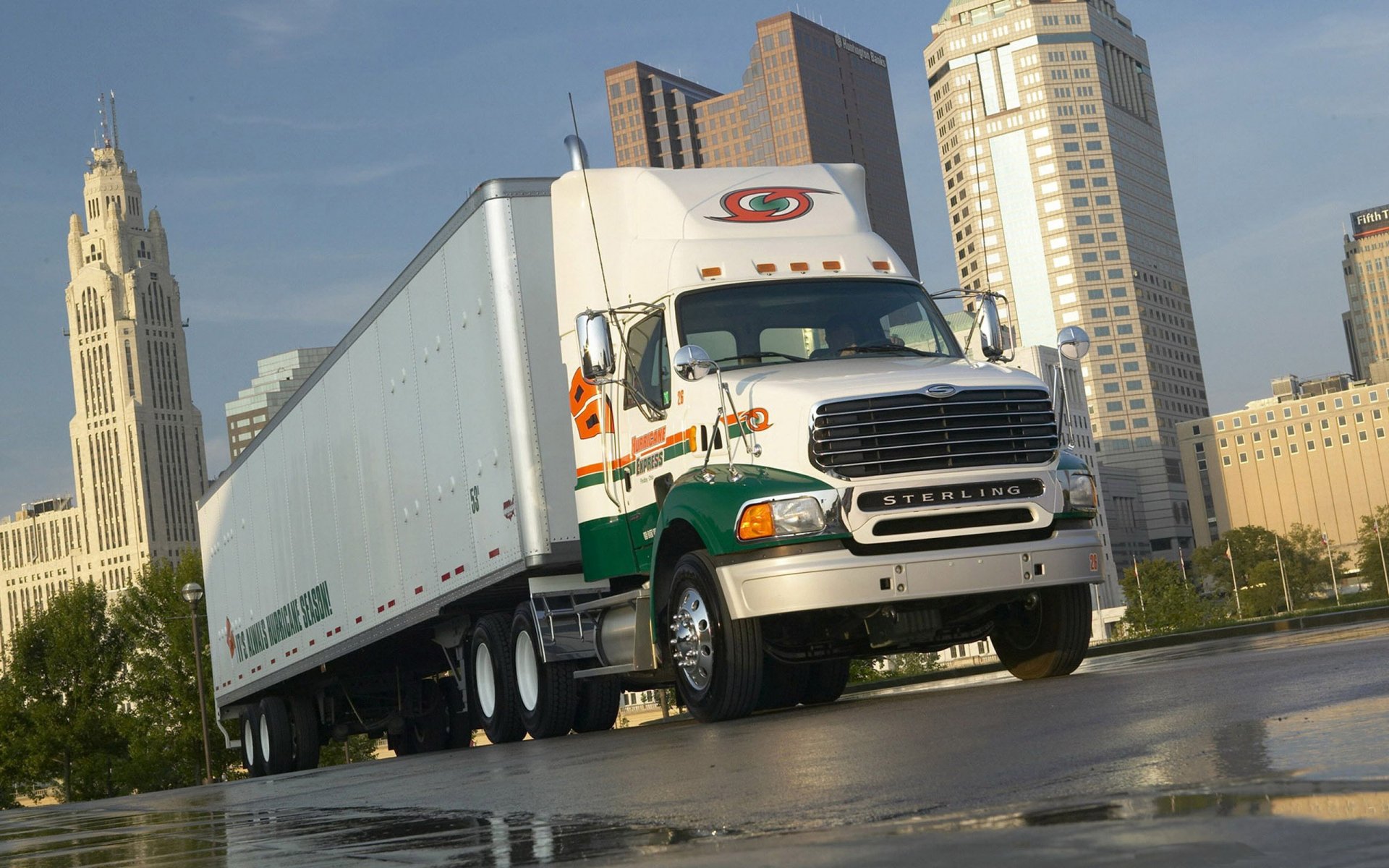 Sterling semi-truck and trailer on wet pavement with downtown skyline reflected in puddles, presented as a 2K Quad HD PC desktop wallpaper.