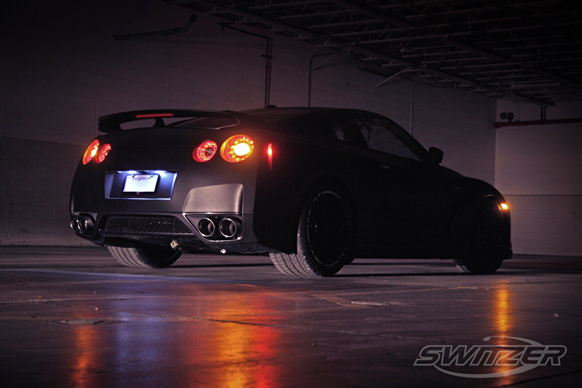 HD PC desktop wallpaper: rear three-quarter view of a black Nissan GT-R parked in a dimly lit garage, glowing taillights and reflections on wet concrete.