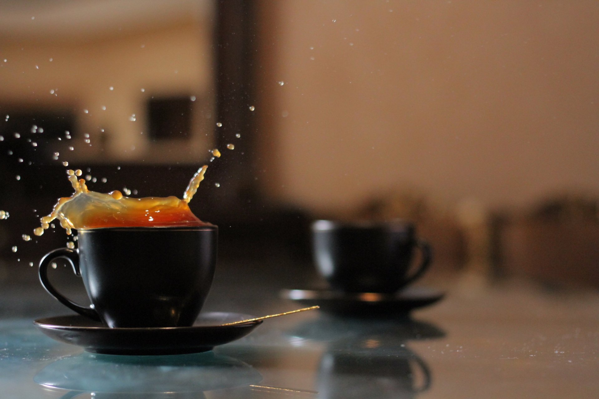 HD desktop wallpaper featuring two black coffee cups with one cup's coffee splashing, set against a softly blurred warm background.