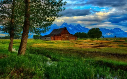 A serene HD wallpaper featuring a rustic barn amid lush green fields and majestic mountains under a dramatic sky, blending man-made charm with natural beauty.