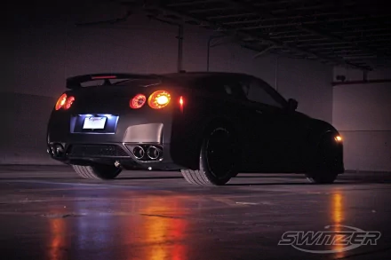 HD PC desktop wallpaper: rear three-quarter view of a black Nissan GT-R parked in a dimly lit garage, glowing taillights and reflections on wet concrete.