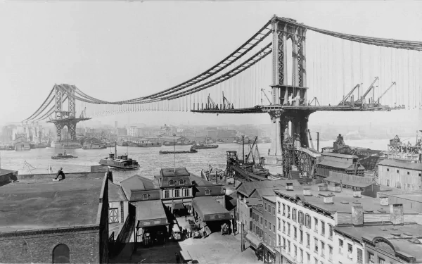 Black and white HD wallpaper showing the Manhattan Bridge spanning a busy river with boats and industrial buildings in the foreground.