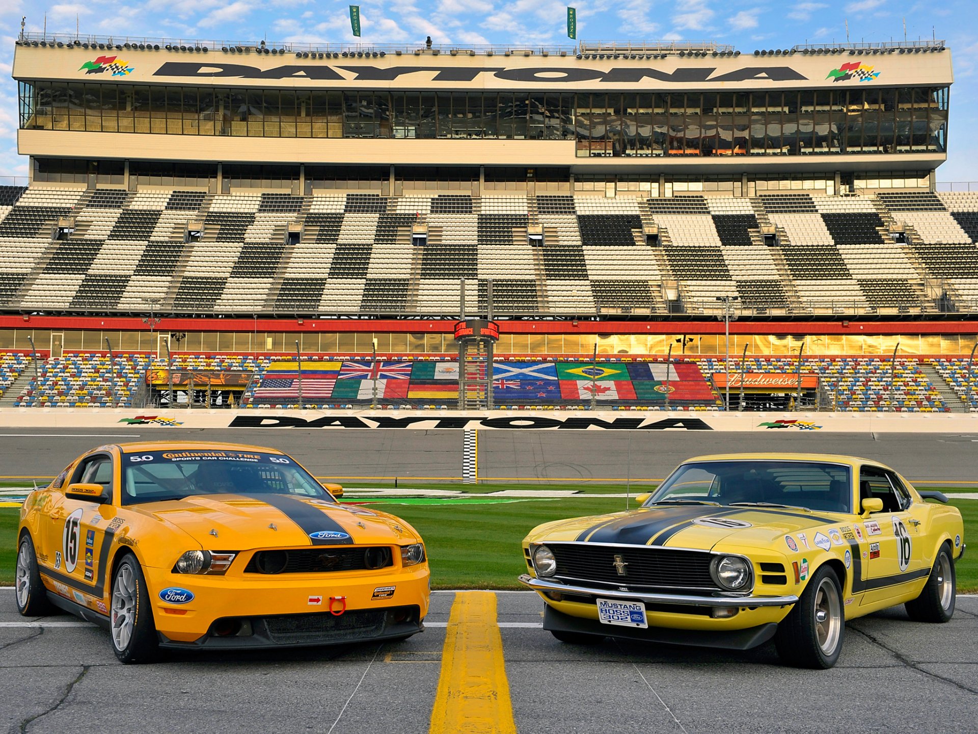 Two Ford Mustang Boss 302 cars, one orange and one yellow, positioned side by side on a racetrack with empty grandstands in the background.