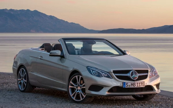 A sleek silver Mercedes-Benz E-Class convertible parked by the water, showcasing its elegant design against a scenic backdrop of mountains and water.