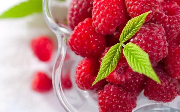 Close-up of vibrant raspberries with green leaves, beautifully arranged in a glass bowl, presented as a 4K Ultra HD desktop wallpaper and background.