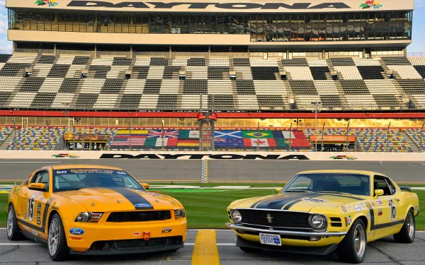 Two Ford Mustang Boss 302 cars, one orange and one yellow, positioned side by side on a racetrack with empty grandstands in the background.
