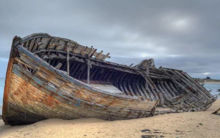 HD desktop wallpaper of an abandoned, weathered wooden shipwreck resting on a sandy shore under a cloudy sky.