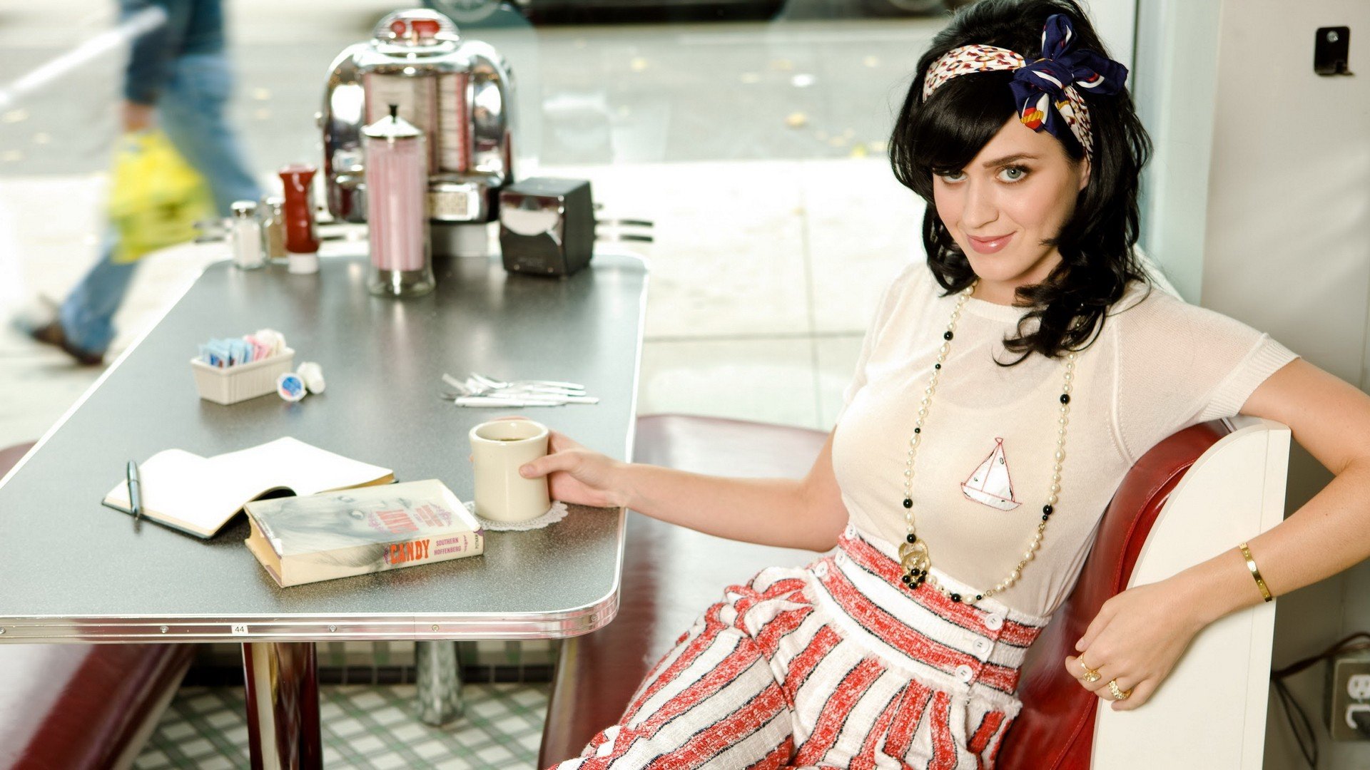 Katy Perry lounges in a retro diner setting, holding a cup of coffee beside a notebook and a novel, captured in this vibrant HD desktop wallpaper.