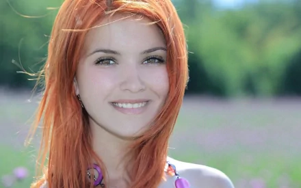 HD desktop wallpaper featuring a woman with red hair smiling brightly, set against a blurred natural background of greenery and flowers.