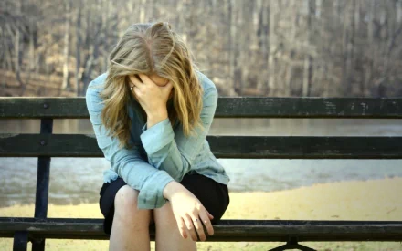 A woman, with her head in her hand, sits on a bench in a forested area. The image evokes a mood of contemplation or sadness and is used as an HD desktop wallpaper and background.